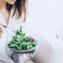 woman in white dress holding silver fork and knife slicing green vegetable on white ceramic bowl